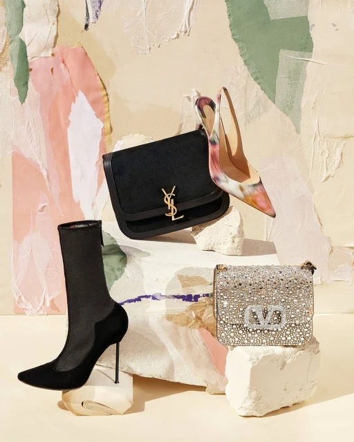 Fashion still life featuring a black YSL purse, a jewel-encrusted Valentino purse, a black stiletto boot, and a colorful stiletto heel. 