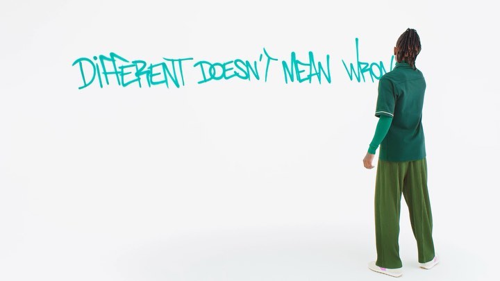 An adult stands in front of a white wall with the turquoise painted slogan: Different Doesn't Mean Wrong, fashion style studio shot.