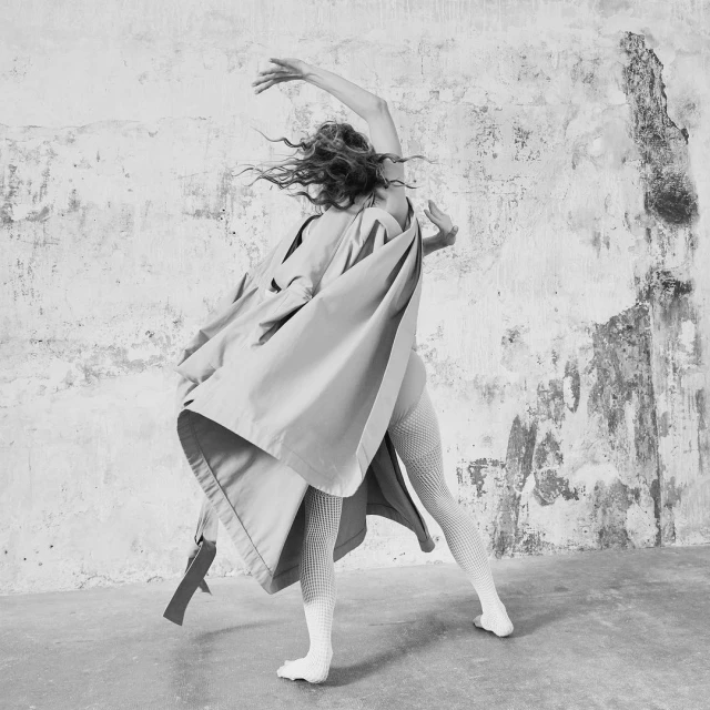 Black and white photograph features a dancer in mid-motion, wearing a coat and textured tights against a worn wall backdrop.