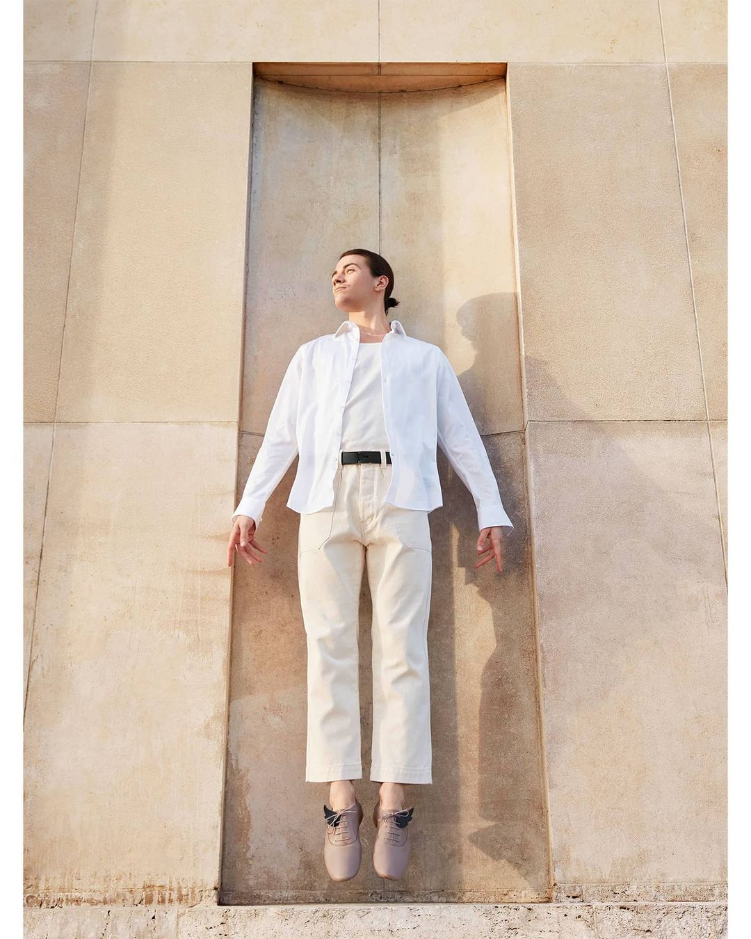 Man in white shirt and beige pants appears suspended in a carved wall opening outdoors, bathed in warm sunlight.
