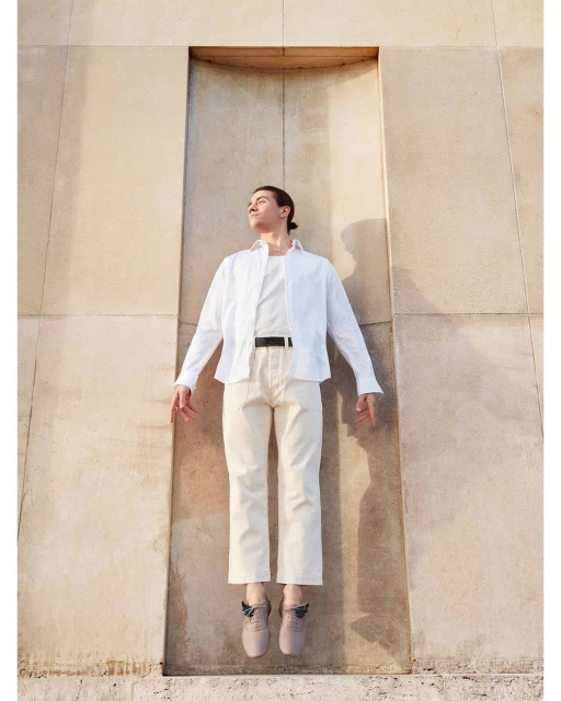 Man in white shirt and beige pants appears suspended in a carved wall opening outdoors, bathed in warm sunlight.