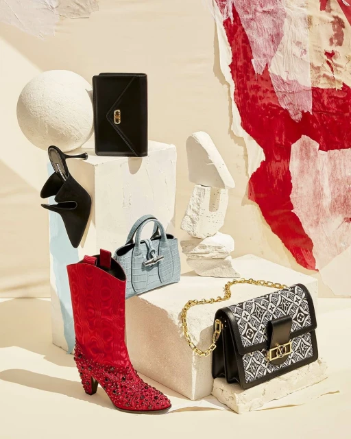 Fashion still life featuring an assortment of designer handbags, a pair of heeled mules, and a western-style boot on white textured blocks.