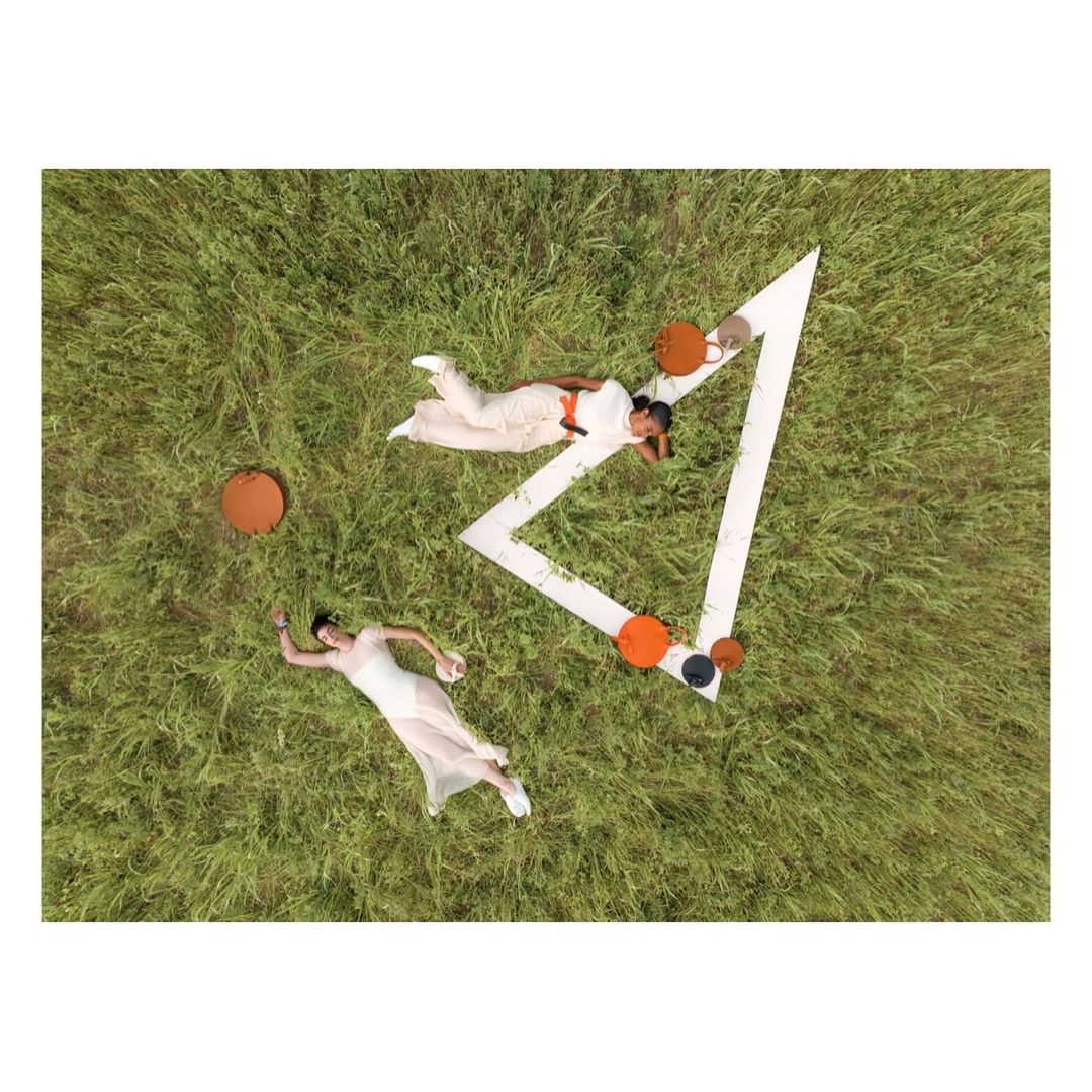 Aerial view of two women in white outfits relaxing on grassy field near geometric set piece with orange and black accent furniture.