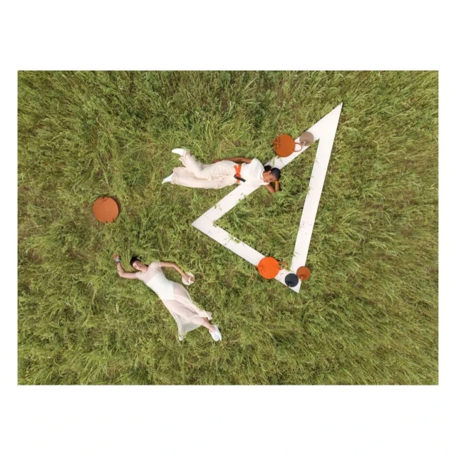 Aerial view of two women in white outfits relaxing on grassy field near geometric set piece with orange and black accent furniture.