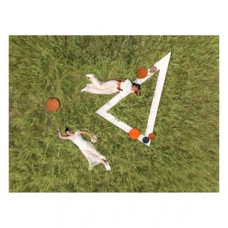 Aerial view of two women in white outfits relaxing on grassy field near geometric set piece with orange and black accent furniture.