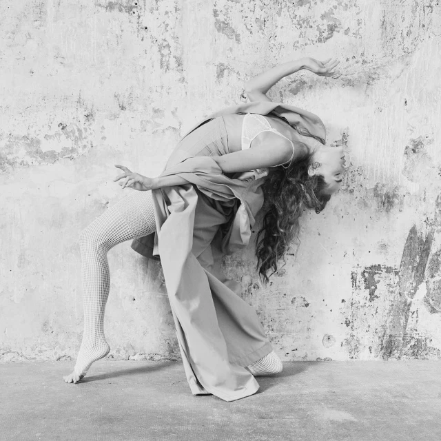 Black and white portrait of a woman arching back with flowing pants and textured tights, against an old distressed wall.