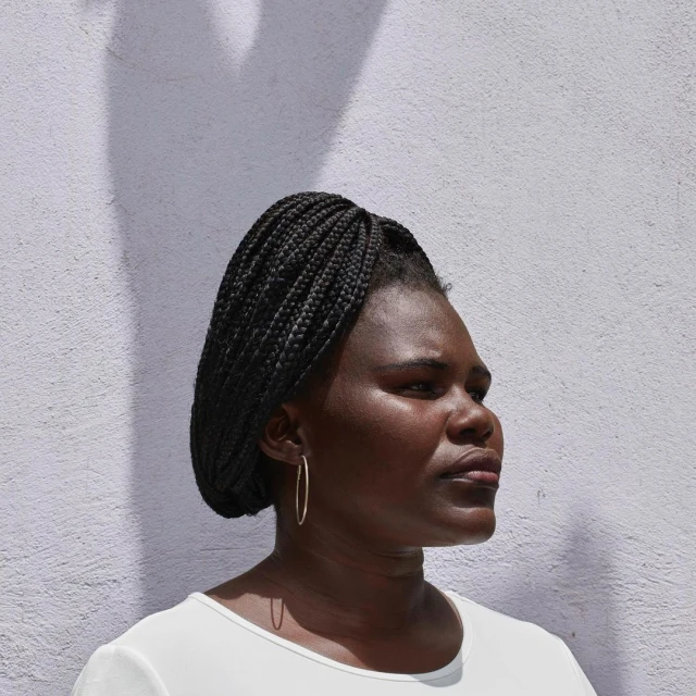 Portrait of a dark-skinned woman with braided hair looking off to the side in front of a mottled white wall with shadow.