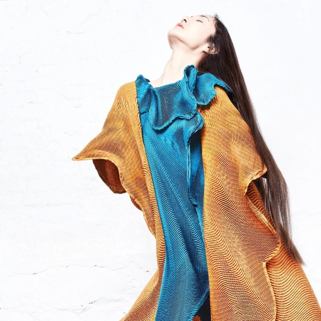 Adult woman with long brown hair poses wearing a gold and turquoise textured dress against a white textured wall, studio lighting.