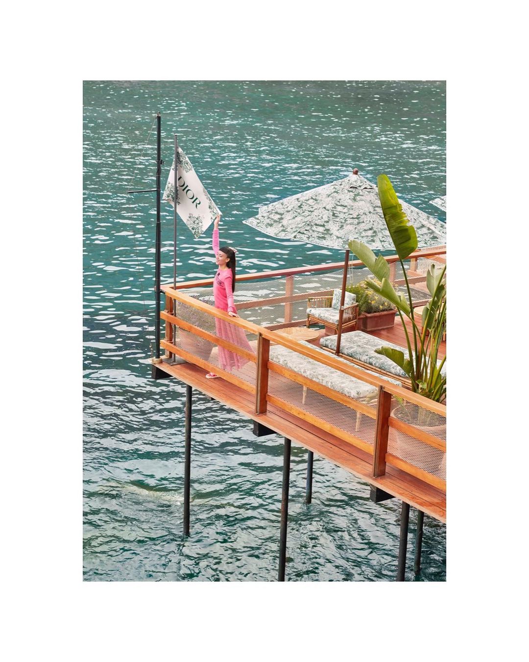 An adult woman stands on a wooden platform holding up a flag with 'DIOR' visible, ocean water and green foliage in the background.