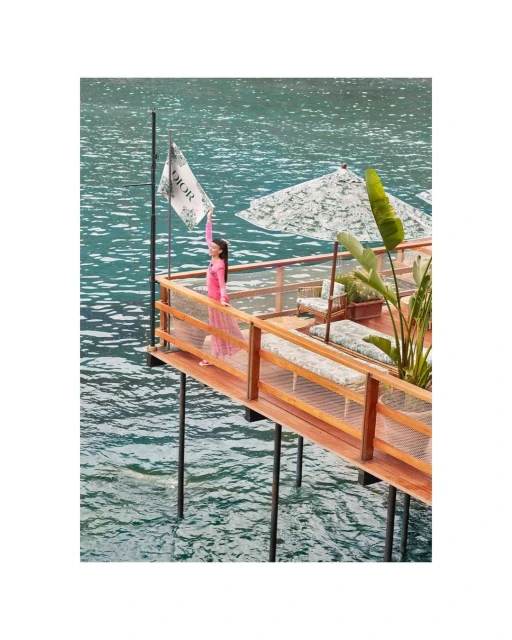 An adult woman stands on a wooden platform holding up a flag with 'DIOR' visible, ocean water and green foliage in the background.