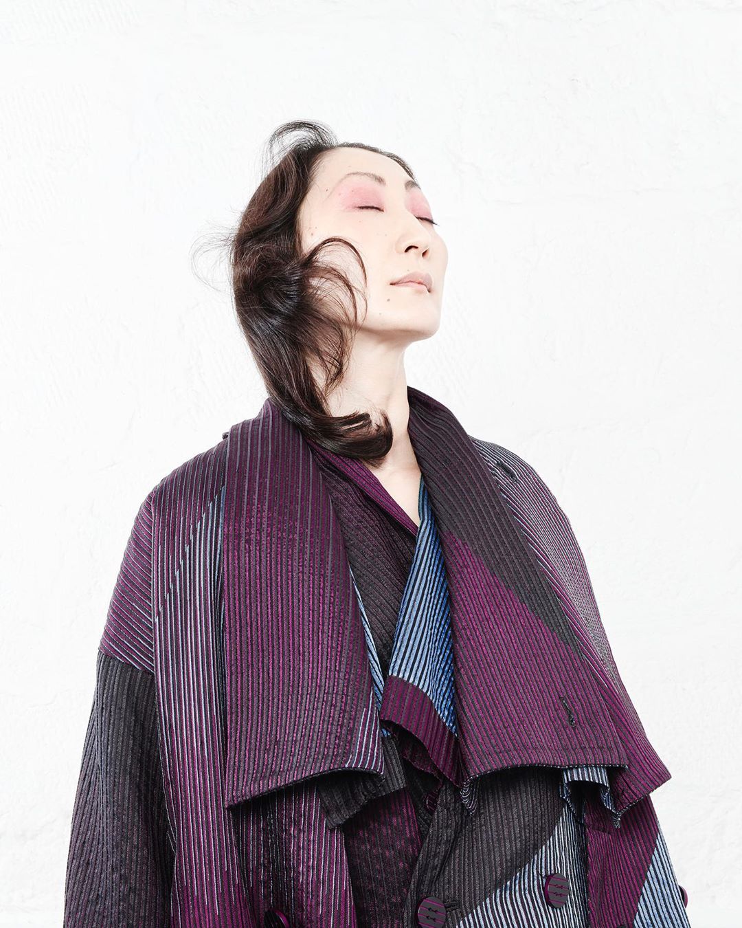 Adult woman in a patterned coat, posing indoors against a white wall with eyes closed, illuminated by soft, diffused lighting.