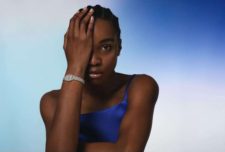 Portrait of a young African-descent woman with hand partially covering face, wearing a blue top and a watch, against a light blue backdrop.
