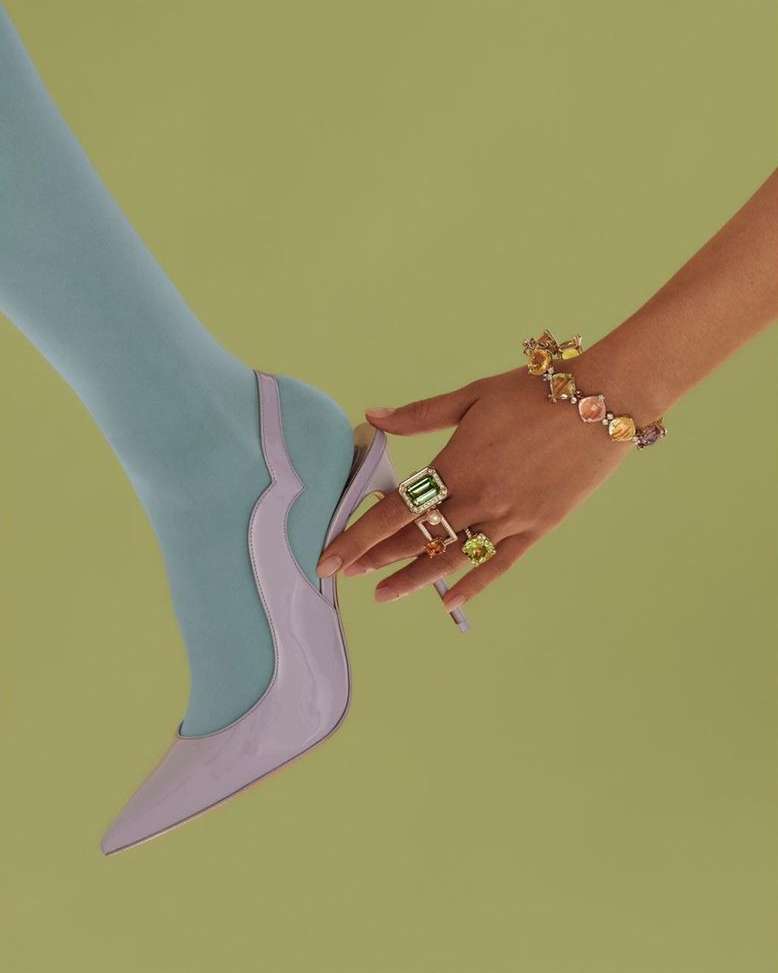 Close-up of a woman's leg in blue tights, juxtaposed with a hand wearing a bracelet and several rings against a lime green background.