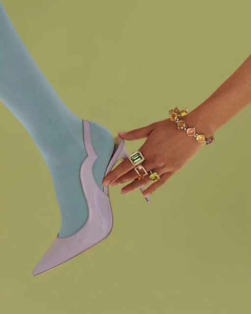 Close-up of a woman's leg in blue tights, juxtaposed with a hand wearing a bracelet and several rings against a lime green background.
