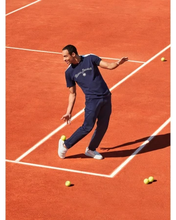 A male tennis player in a blue t-shirt and pants stands on a red clay court surrounded by scattered tennis balls in bright sunlight.