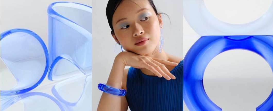 Asian woman with blue eyeshadow and transparent jewelry poses in a studio with curved translucent objects in a beauty fashion shoot.