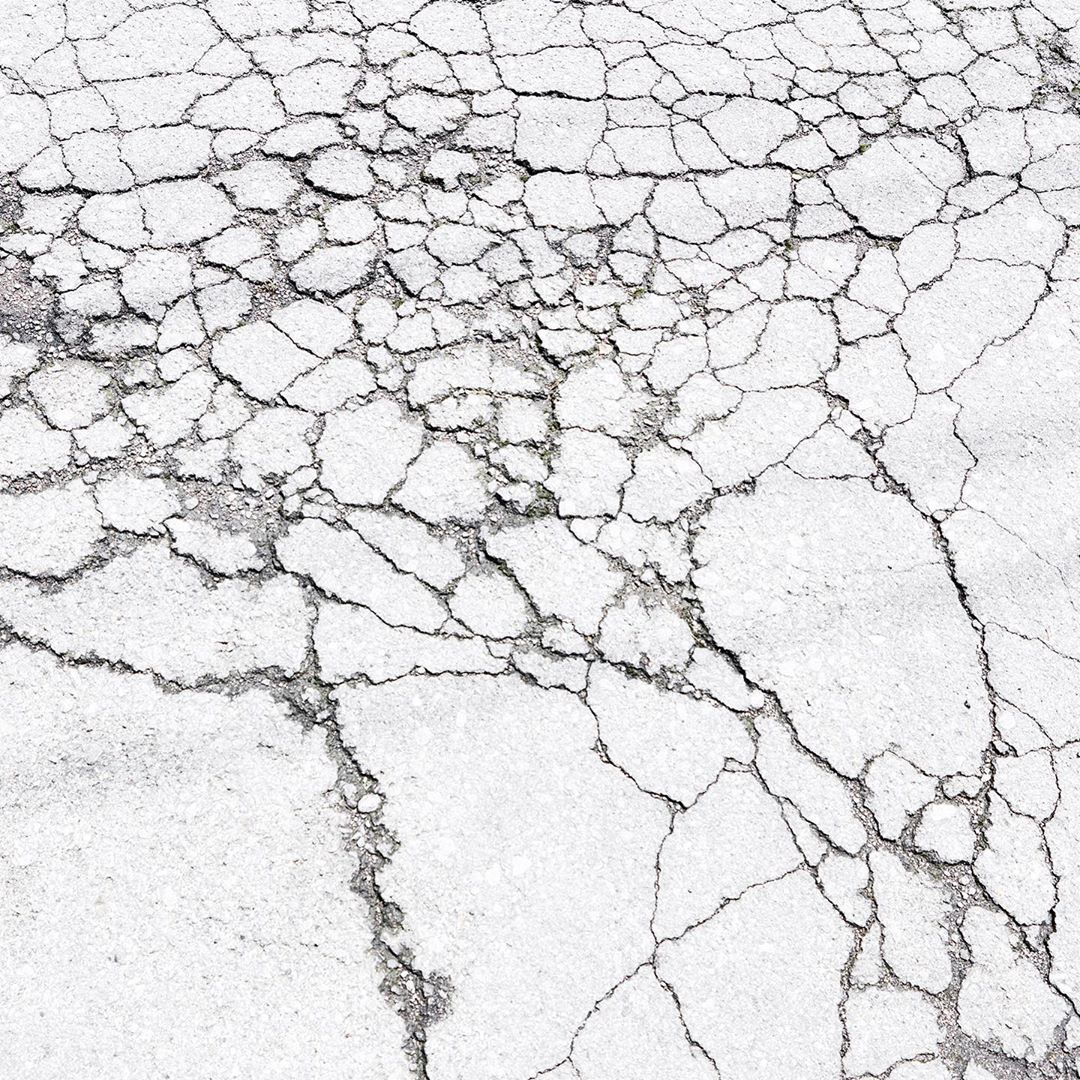 Close-up image of cracked, grey and white asphalt surface featuring complex network of fissures and textured road damage.