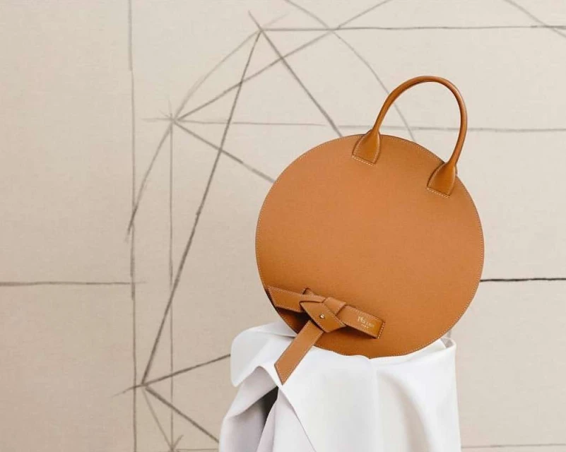 Tan leather circle handbag with round handles resting on draped white fabric in a minimalist setting with linear geometric background.