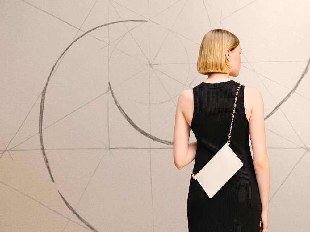 Back view of a blonde woman wearing a black dress, with a white clutch bag on a chain strap, against a wall with abstract art.