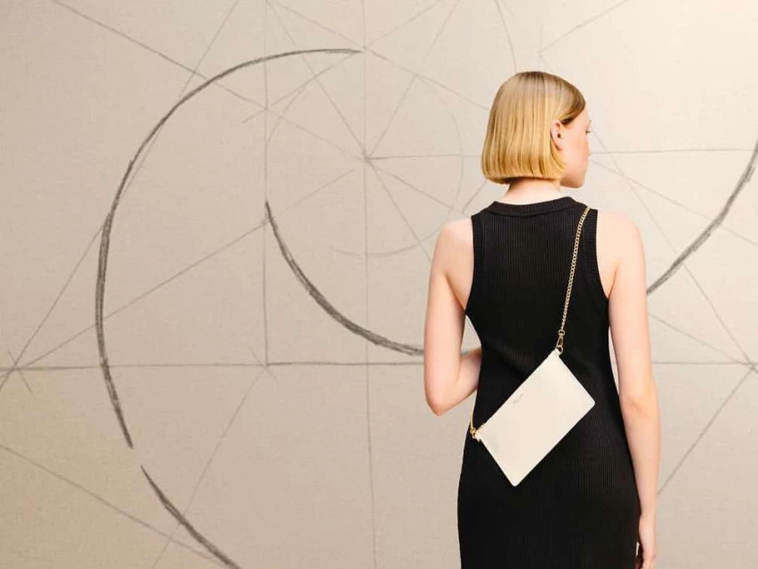 Back view of a blonde woman wearing a black dress, with a white clutch bag on a chain strap, against a wall with abstract art.