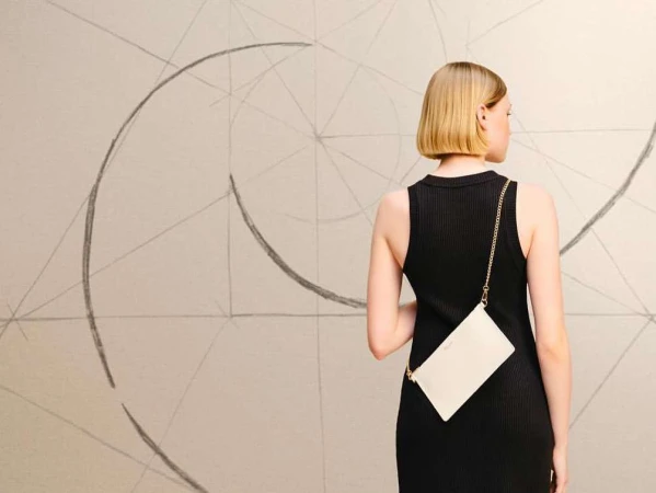 Back view of a blonde woman wearing a black dress, with a white clutch bag on a chain strap, against a wall with abstract art.