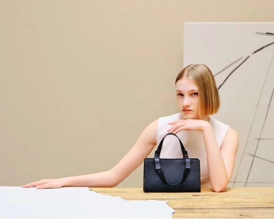 Blonde woman wearing a cream top posing behind a dark handbag on a wooden table with an abstract art piece in the background