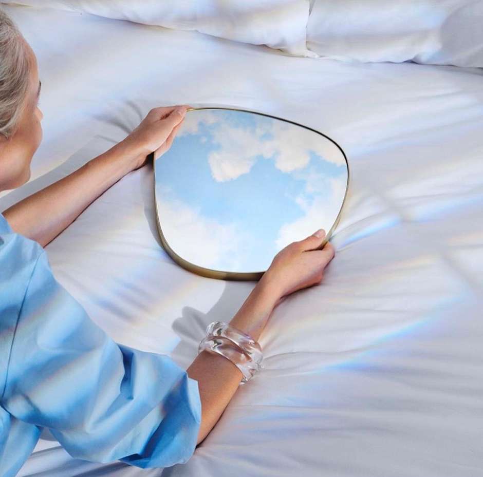 Silver-haired woman lying on a bed, holding a gold framed square mirror reflecting the sky and clouds, wearing a translucent bracelet.