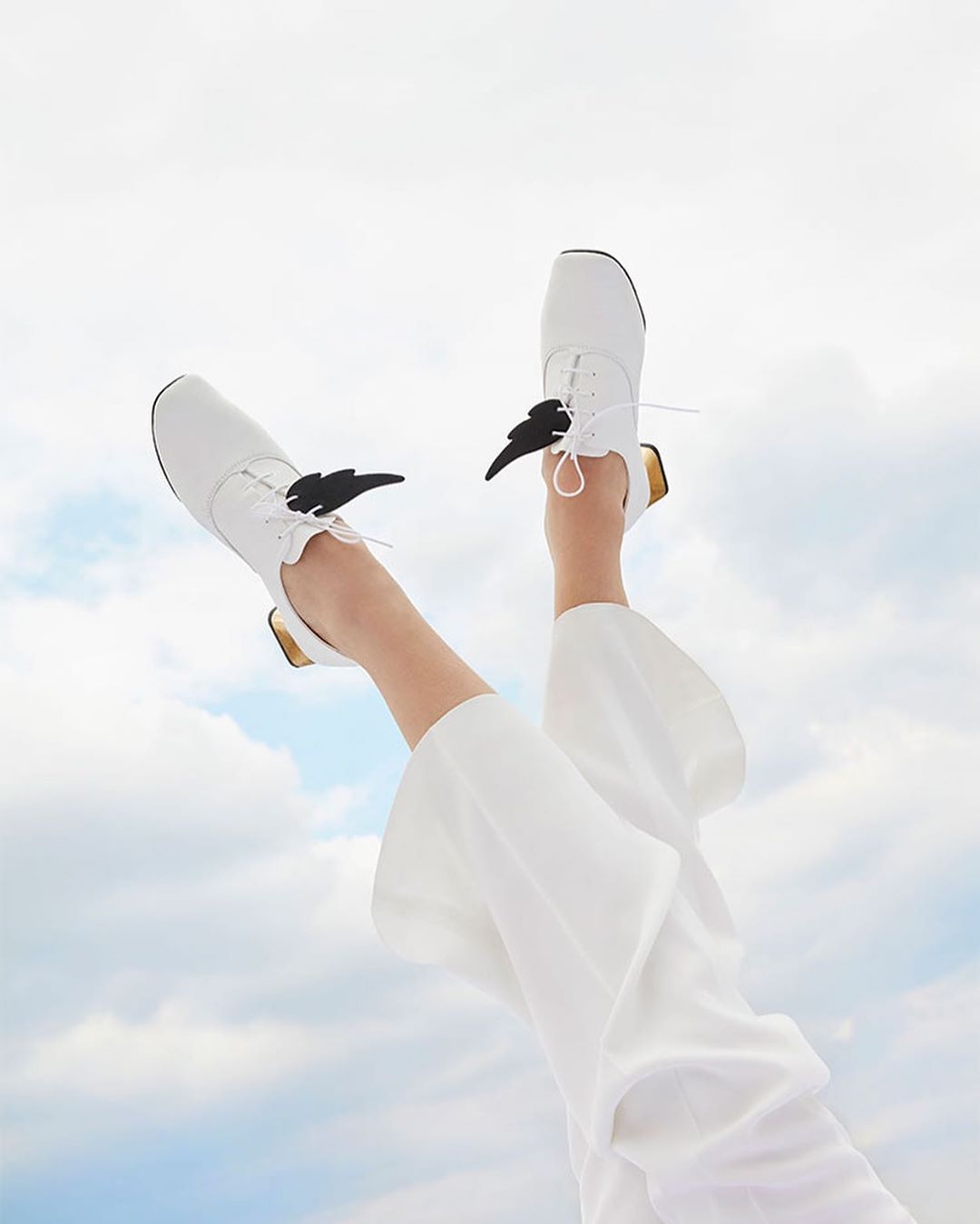 Legs and feet wearing white shoes with black wings pointing up against a cloudy sky. Bright, aerial fashion footwear.