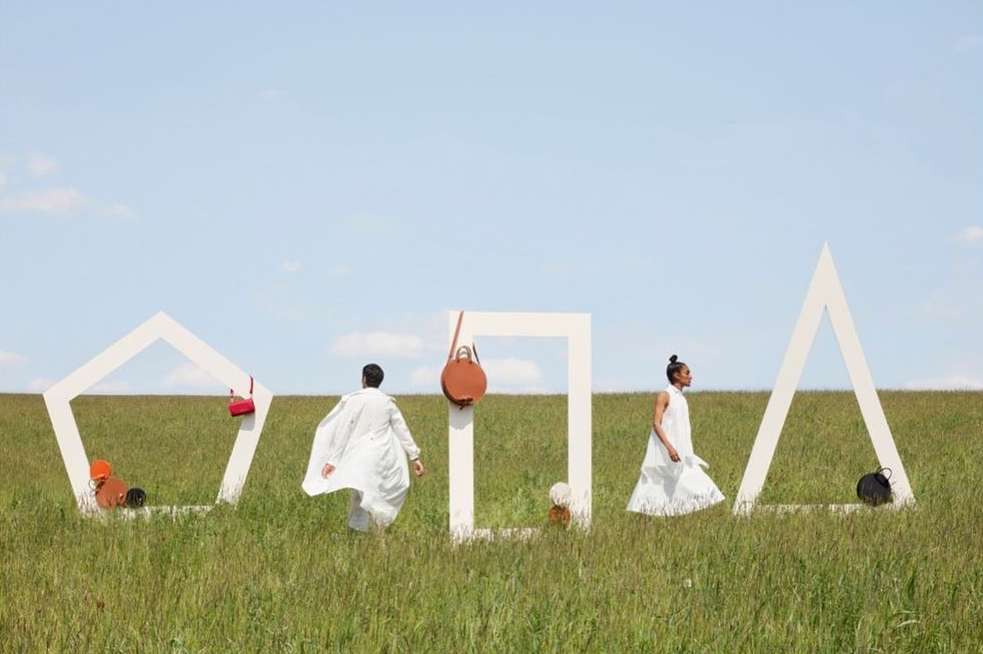 Fashion editorial on a grassy field with two people in white outfits among white geometric shapes and colorful handbags hanging within.