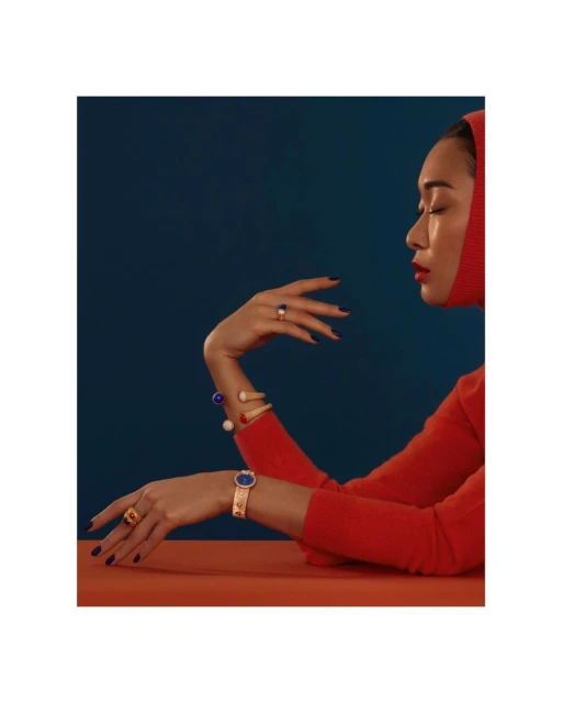 Profile of a woman in red clothing and jewelry, including rings and a watch, with a dark blue background, resting her arms on a table.