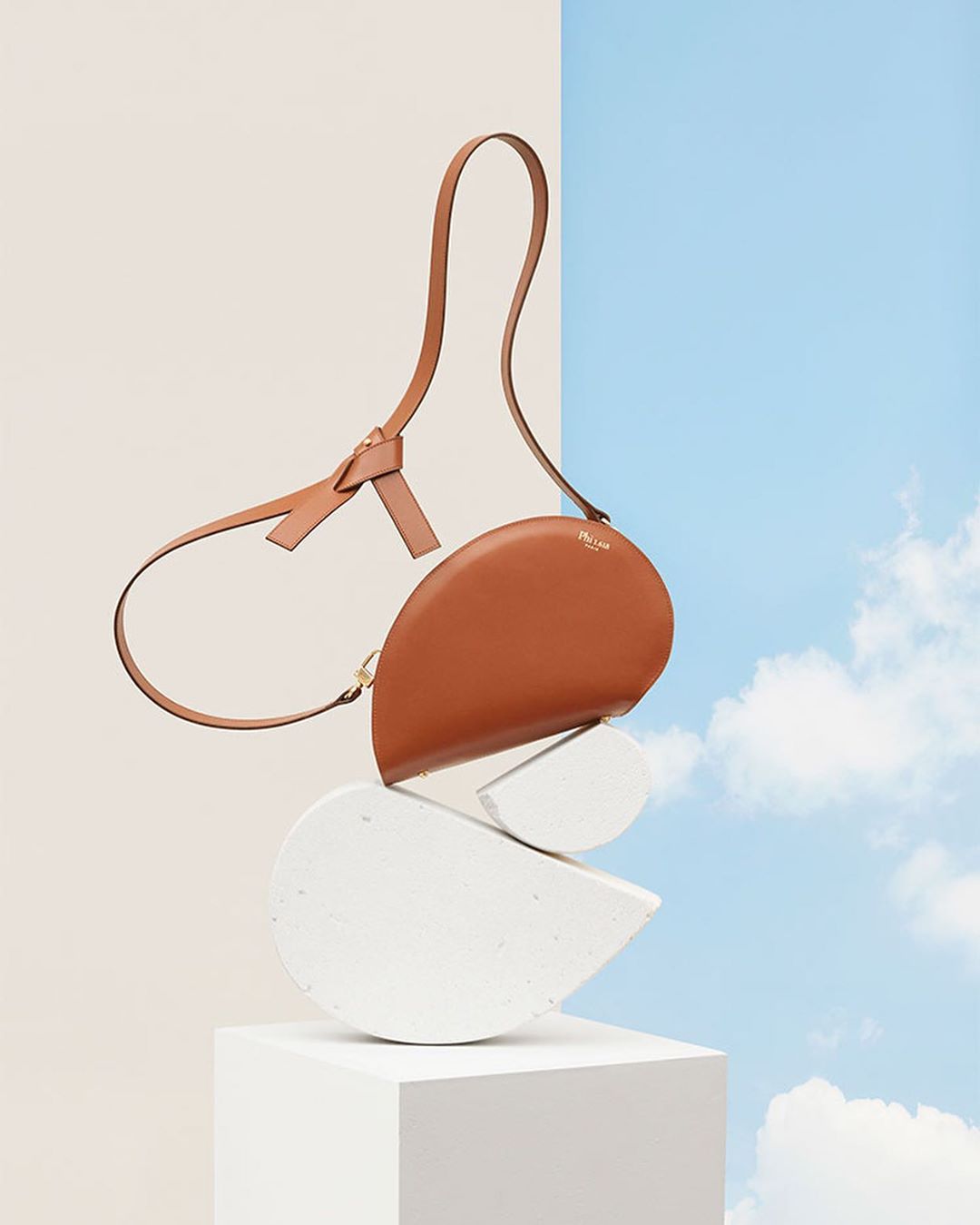 Studio shot of a brown leather purse balancing on stacked white geometric stones against a split beige and sky-blue background.