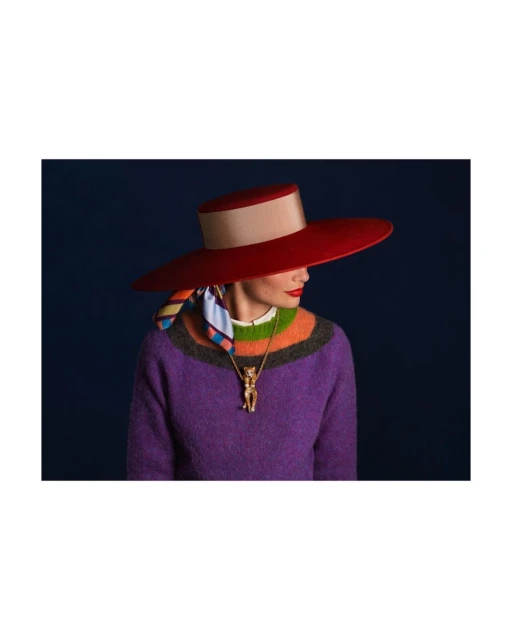 Elegant adult woman wearing a wide-brimmed red hat, multi-colored striped sweater, and a gold cat pendant against a dark blue background.
