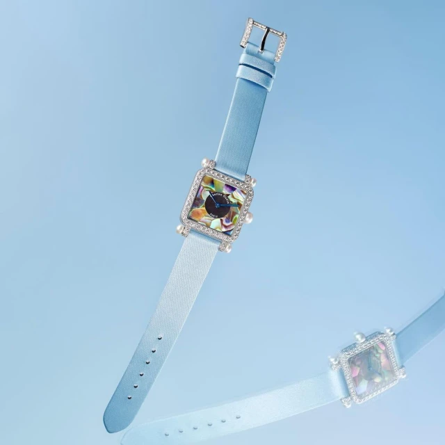 Elegant square wristwatch on a light blue background, showcasing its jeweled bezel and light blue leather strap in a studio product shot.