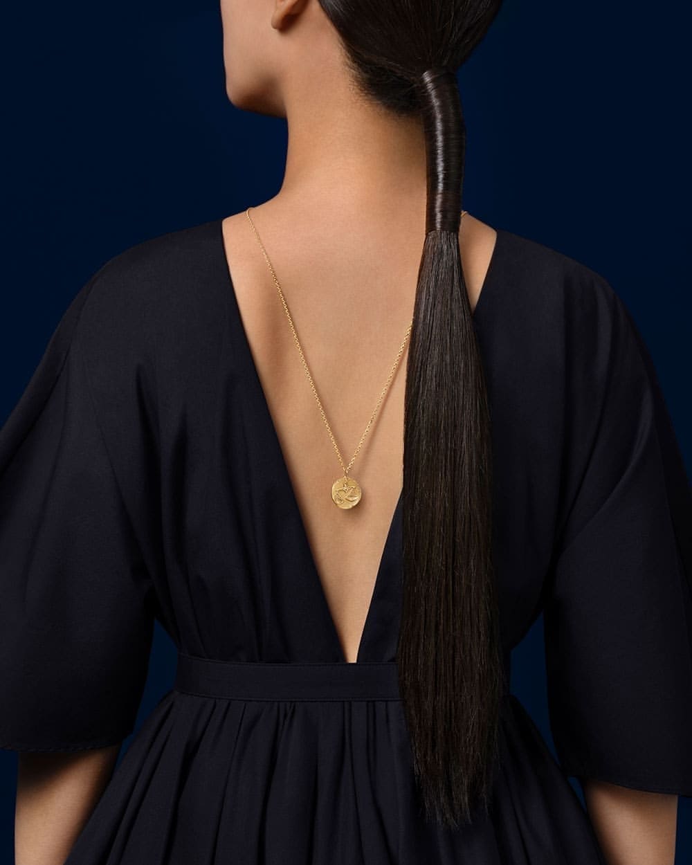Back view of a female model wearing a black dress and gold necklace against a dark blue background in a studio setting.