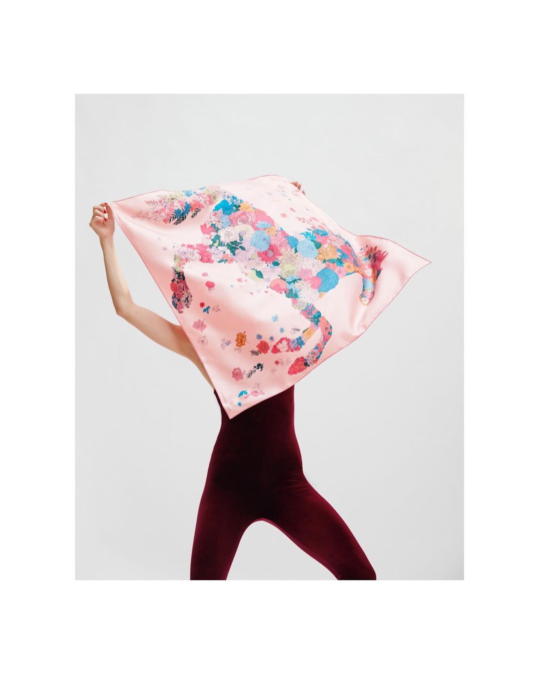 Woman holds a pink silk scarf with a floral horse design against a white backdrop, fashionably showcasing its vibrant details.
