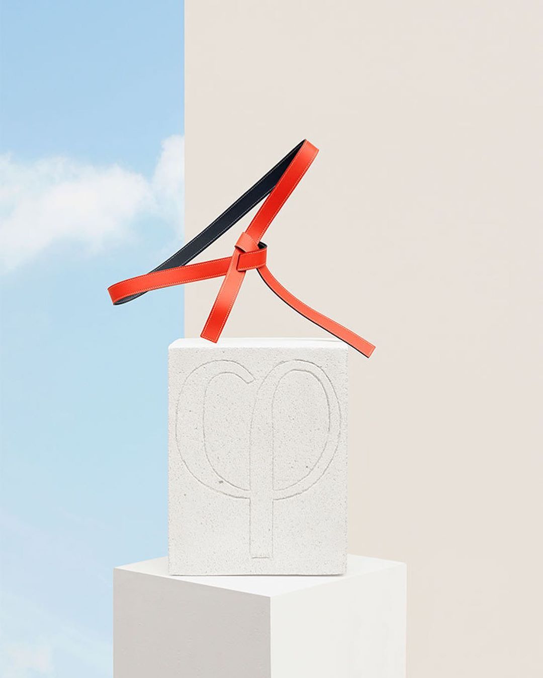 Modern still life of a tied leather strap on a logo-embossed concrete block against a split sky blue and beige background in a studio.