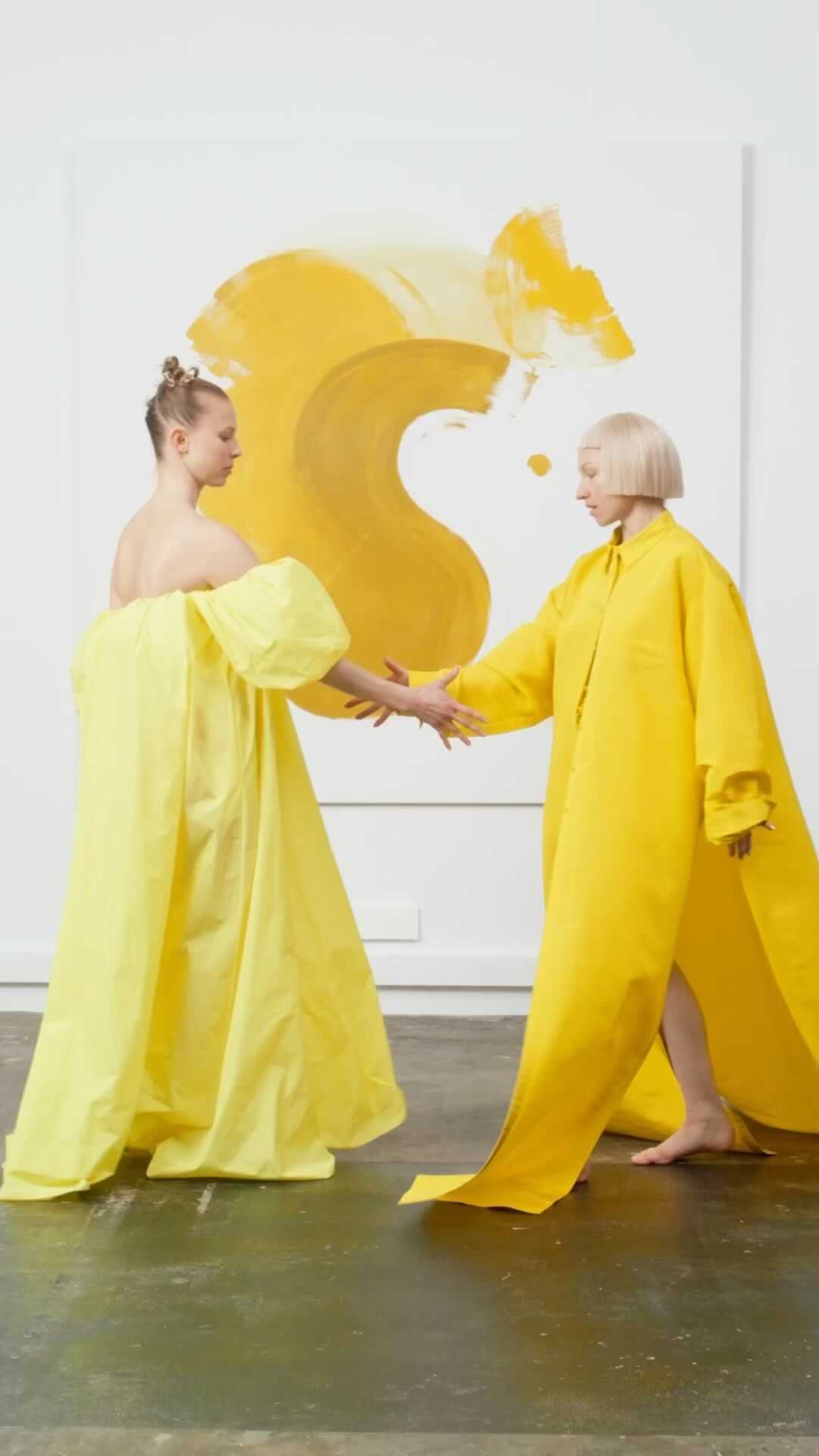 Two women in bright yellow gowns interact in a bright white room with minimal decor and an abstract painting, creating a fashion editorial.