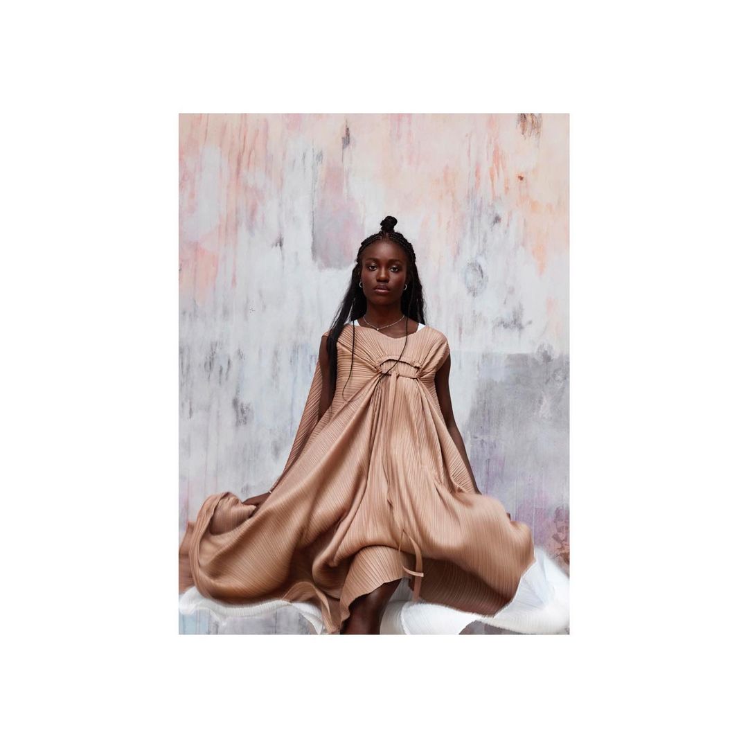 Elegant Black woman poses in a flowing tan dress against a textured backdrop, creating a fashion editorial image.