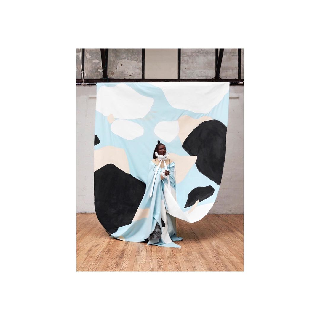 A woman in draped blue and white fabric stands in front of a patterned backdrop in a minimal indoor space with wooden flooring.