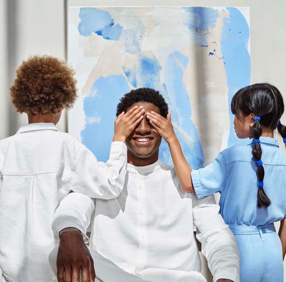 Smiling adult male with dark hair playfully has eyes covered by a young child with afro, as another child with braids looks on.