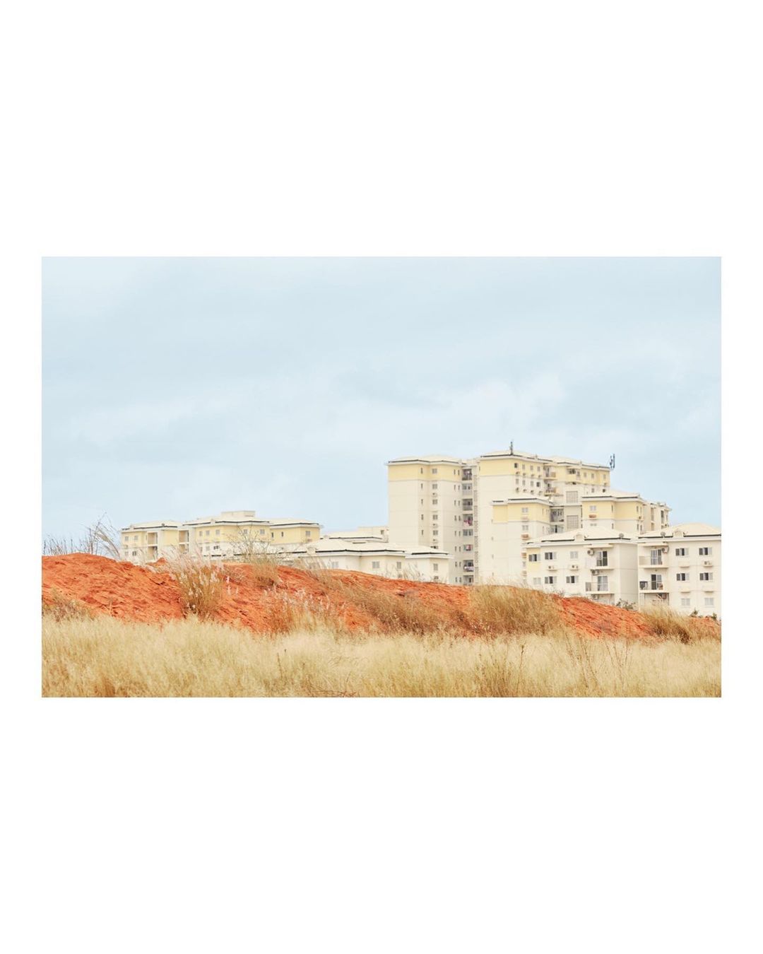 Urban scene featuring apartment buildings with an orange embankment and dry grass in the foreground under a cloudy sky.