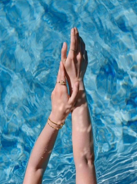 Woman's arms and hands with rings and a bracelet, held above turquoise blue water with rippled surface and bright reflections.