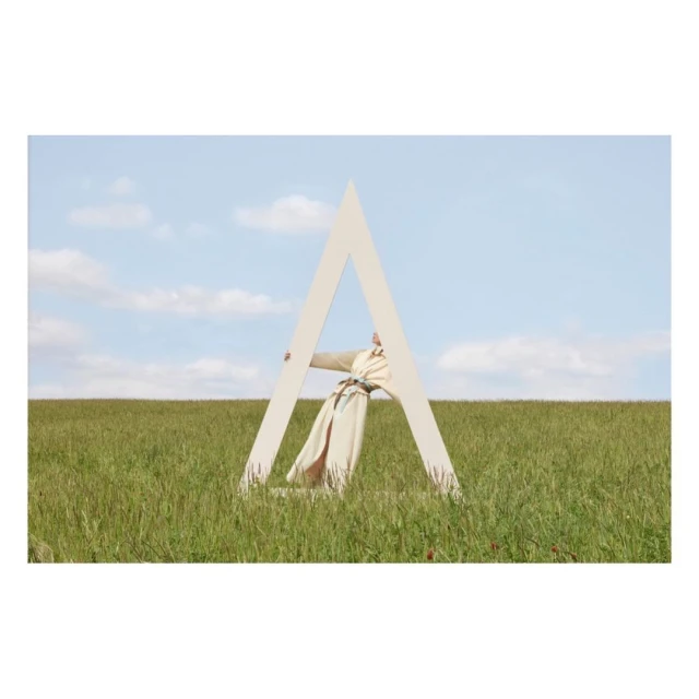 Model in a wheat field standing behind an oversized letter 'A' wearing a beige trench coat and belt, under a blue sky with clouds.