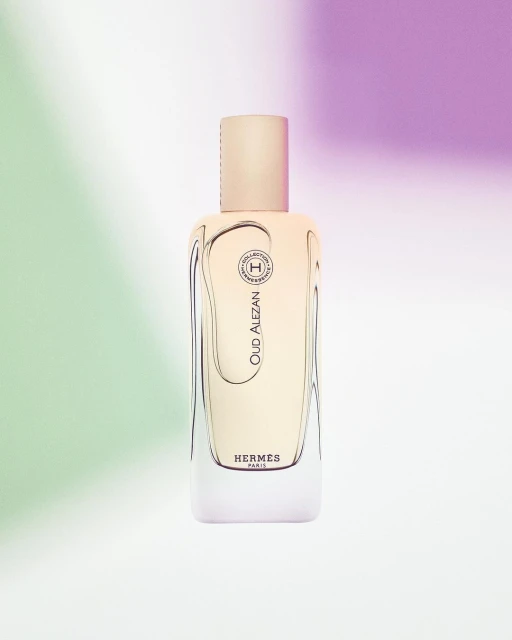 Studio shot of an 'Oud Alezan' perfume bottle by Hermès with pale yellow liquid on a soft white background with pastel green and purple.