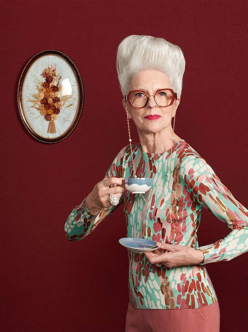Elegant older woman with white bouffant hairstyle wearing glasses and holding a tea cup and saucer in an indoor setting.