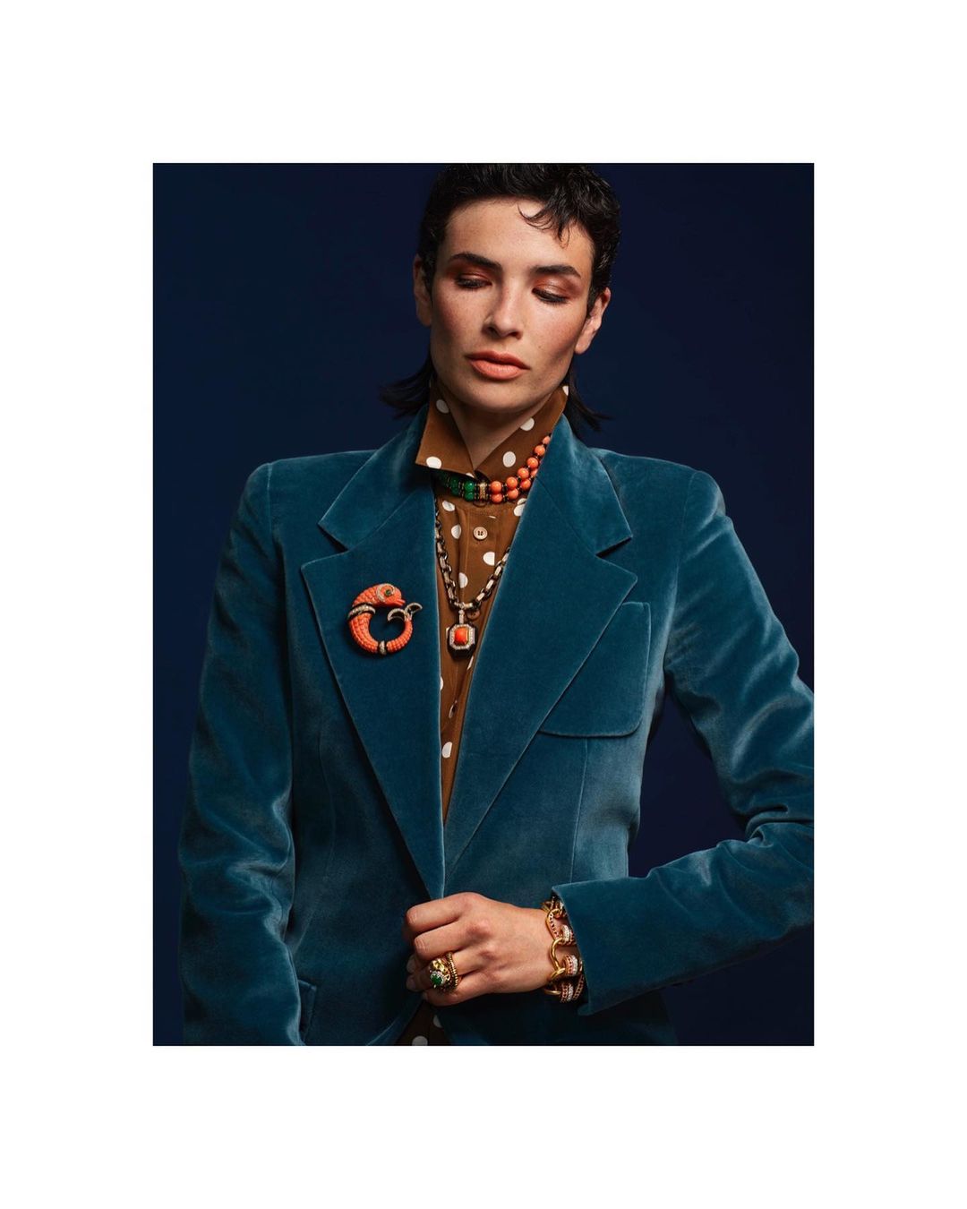 Fashionable woman in a teal velvet blazer and jewelry poses against a navy blue background in a studio setting.