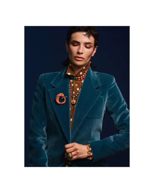 Fashionable woman in a teal velvet blazer and jewelry poses against a navy blue background in a studio setting.