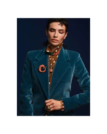 Fashionable woman in a teal velvet blazer and jewelry poses against a navy blue background in a studio setting.