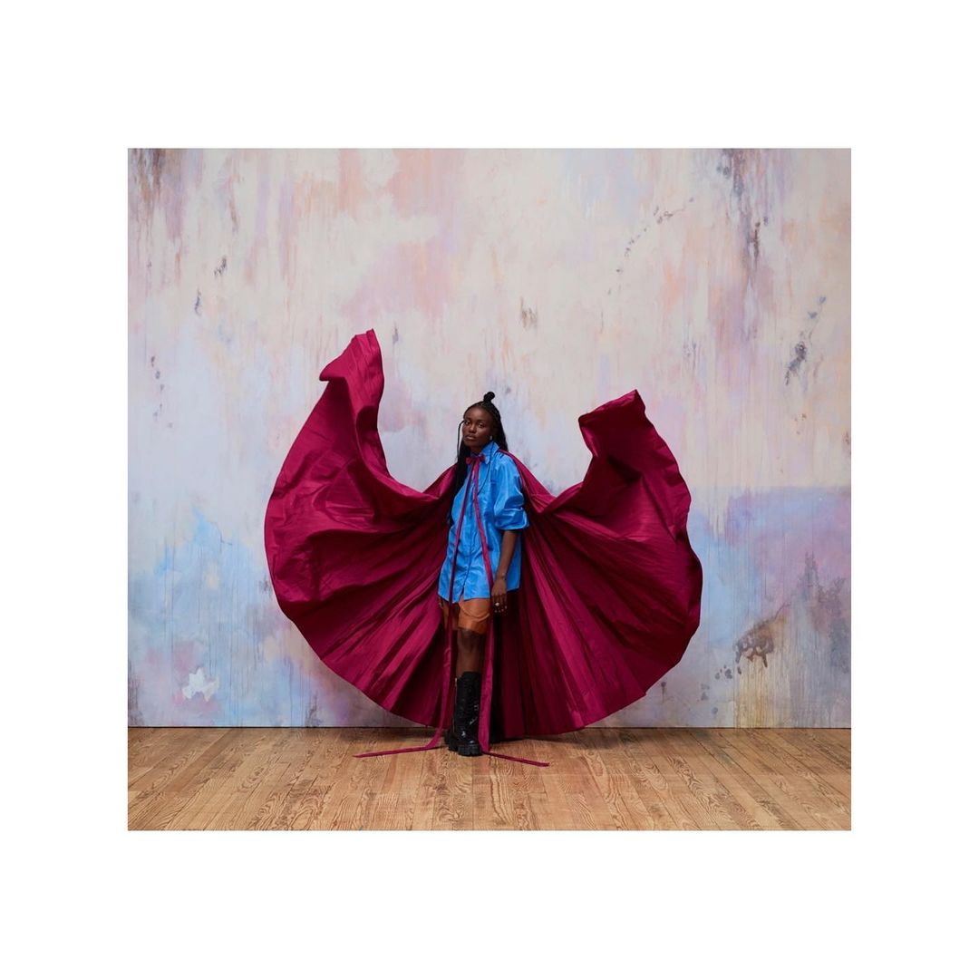 An elegant woman in a blue shirt and flowing magenta fabric poses in a studio setting. She has a modern hairstyle and stands on a wood floor.