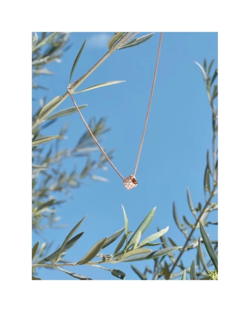 Elegant rose gold necklace with a geometric pendant is suspended against a bright blue sky and olive branches, outdoor fashion jewelry shoot.